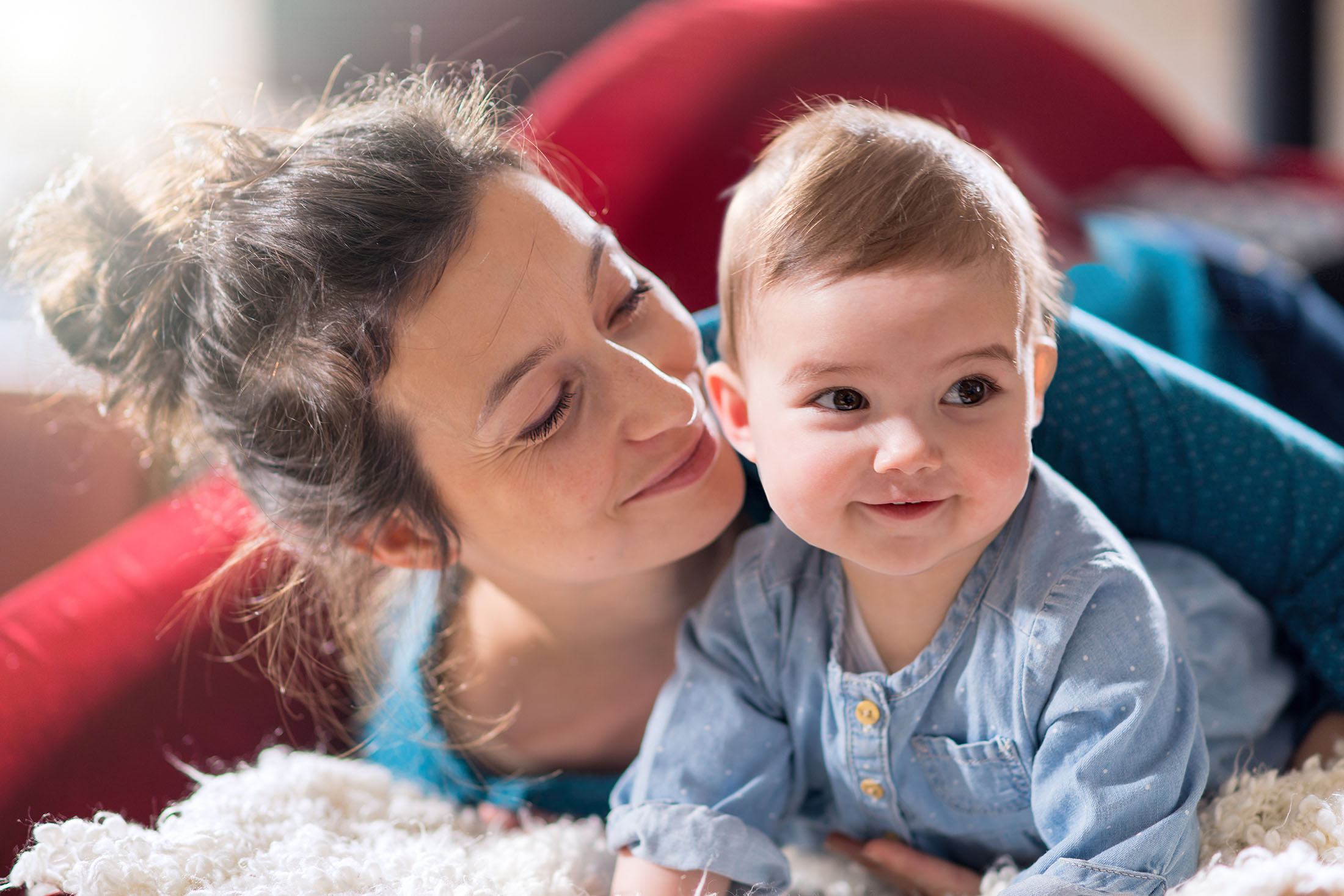 Mother is playing with her cheerful baby on the couch at home. They are looking at camera